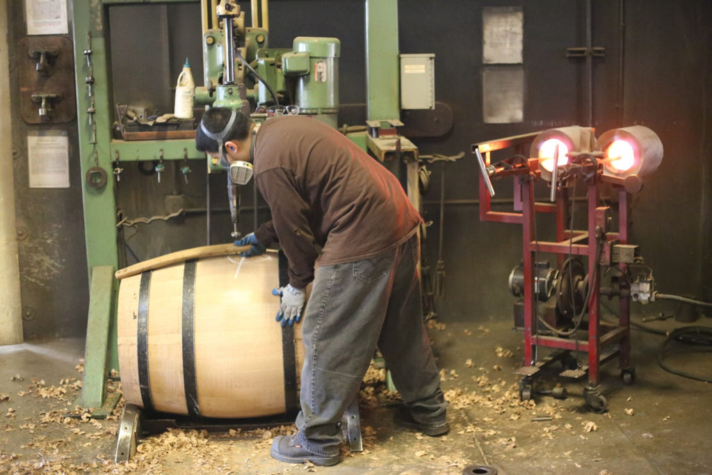 A man in a brown shirt uses a red machine to drill into a barrel, with a white bag visible in the background.