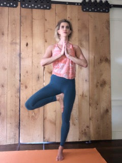 A woman balancing on one leg with hands together, wearing active yoga pants and a top, standing indoors.