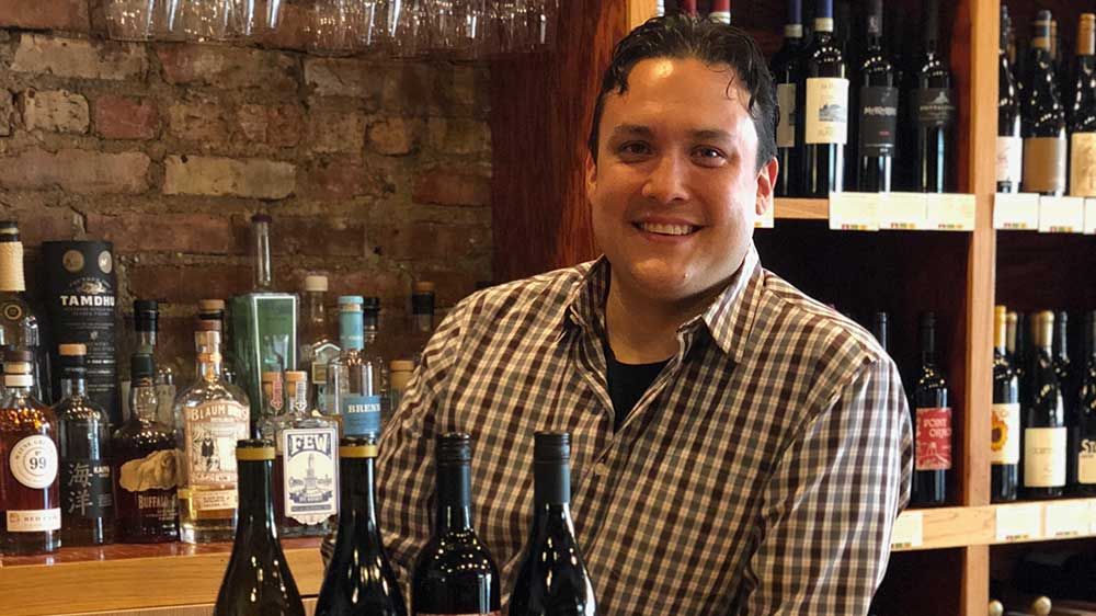 A man smiling at the camera with several close-up bottles of alcoholic beverages visible in the background.