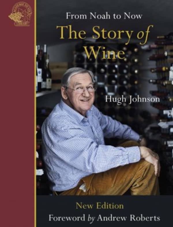 Man in checkered shirt sitting in wine cellar, holding The Story of Wine: From Noah to Now by Hugh Johnson, smiling at the camera.